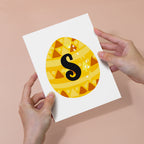 Card with a yellow circle featuring a black letter 'S' held by two hands against a beige background