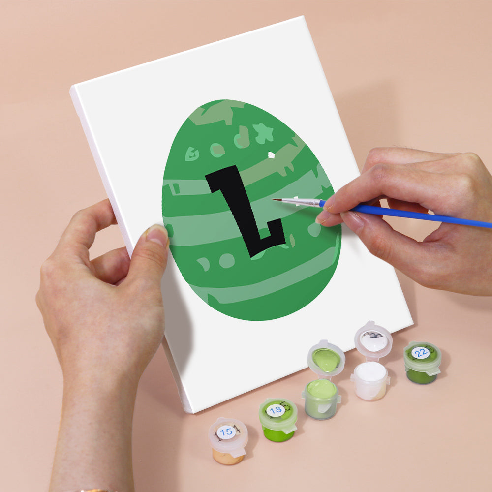 Person painting a green circle with a black letter 'L' on a white card, surrounded by paint containers.
