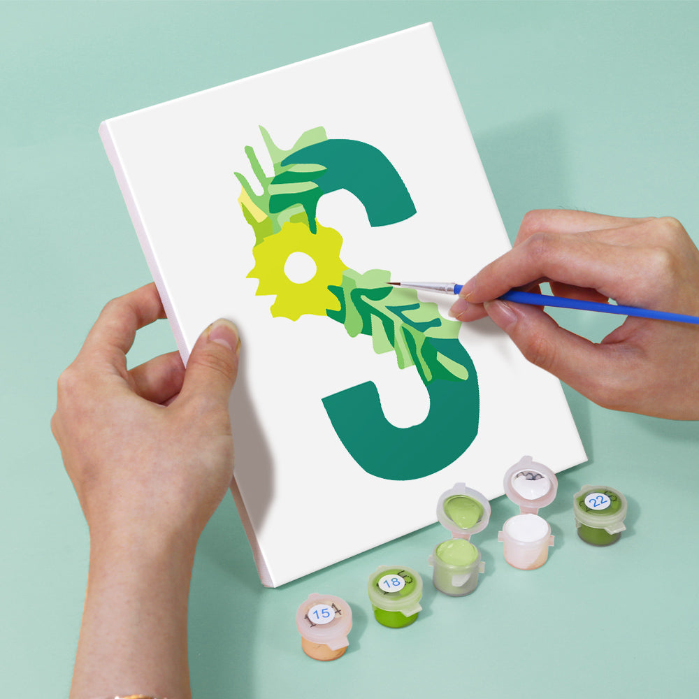 Person painting a green letter 'S' on a white canvas with paint supplies nearby.