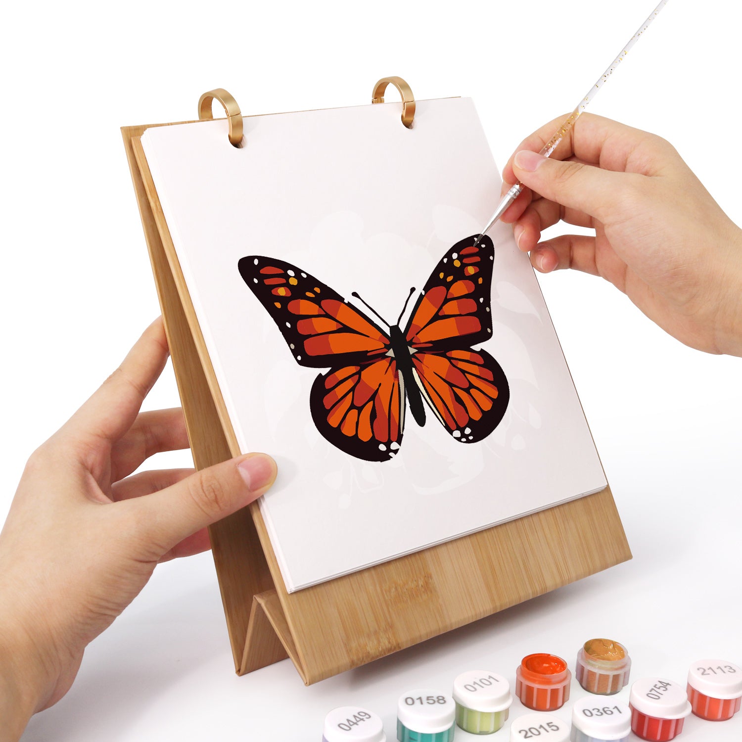 Hand holding a painting of a butterfly on a small easel with art supplies in the background.