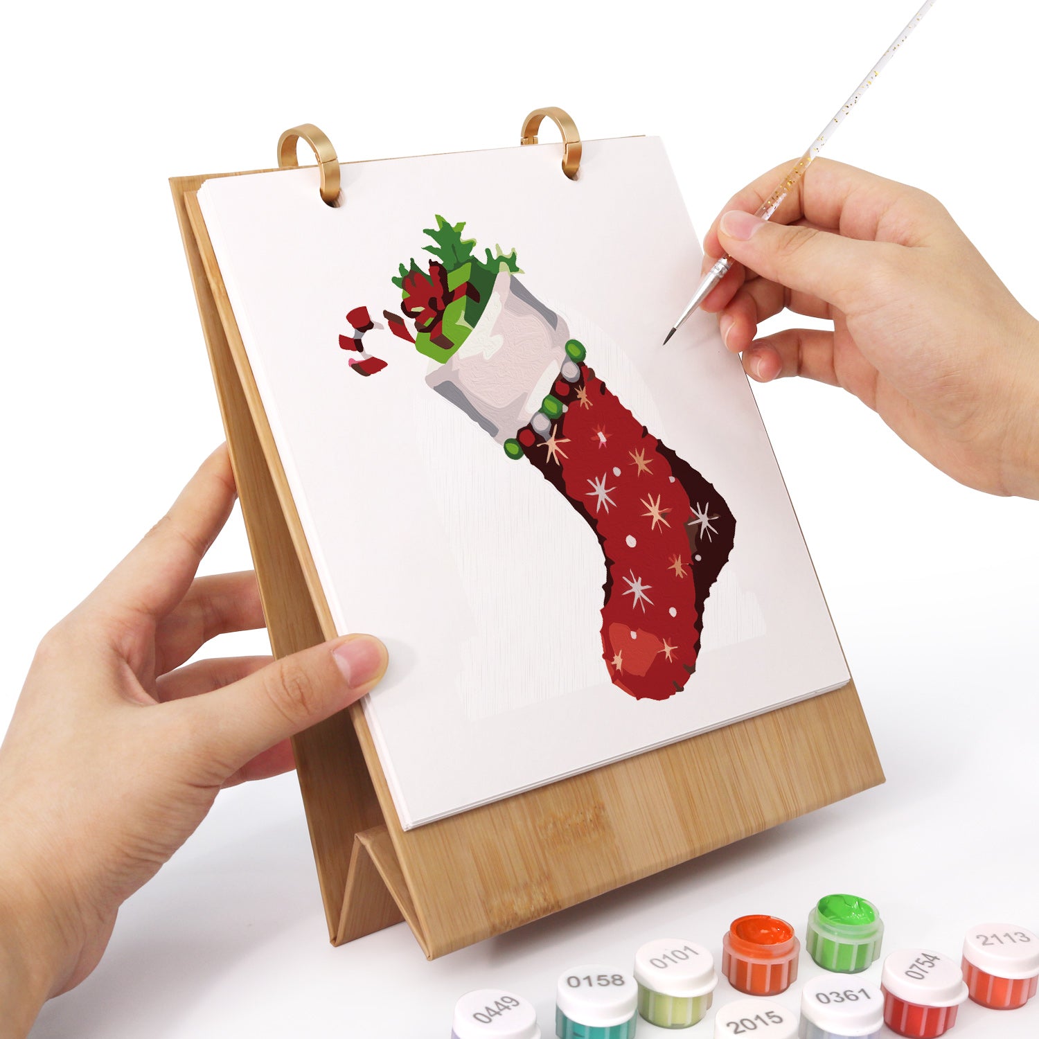 Persona pintando un calcetín con temática navideña en un pequeño caballete con materiales de pintura.