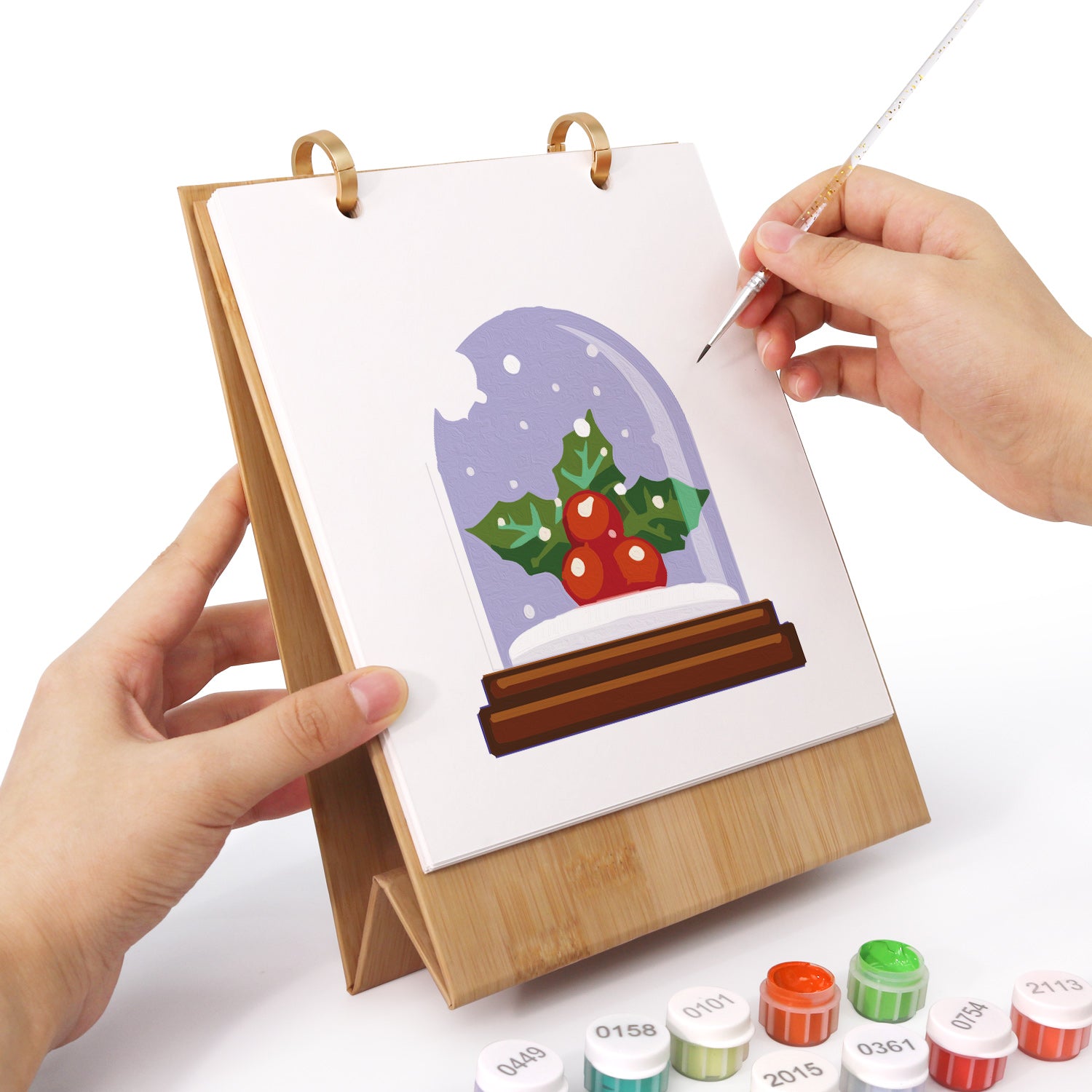 Person painting a snow globe scene on a small easel with paint supplies.