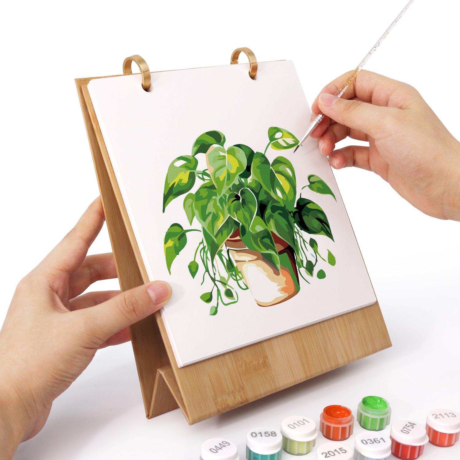 Persona pintando una planta verde sobre un lienzo blanco con un caballete de madera.