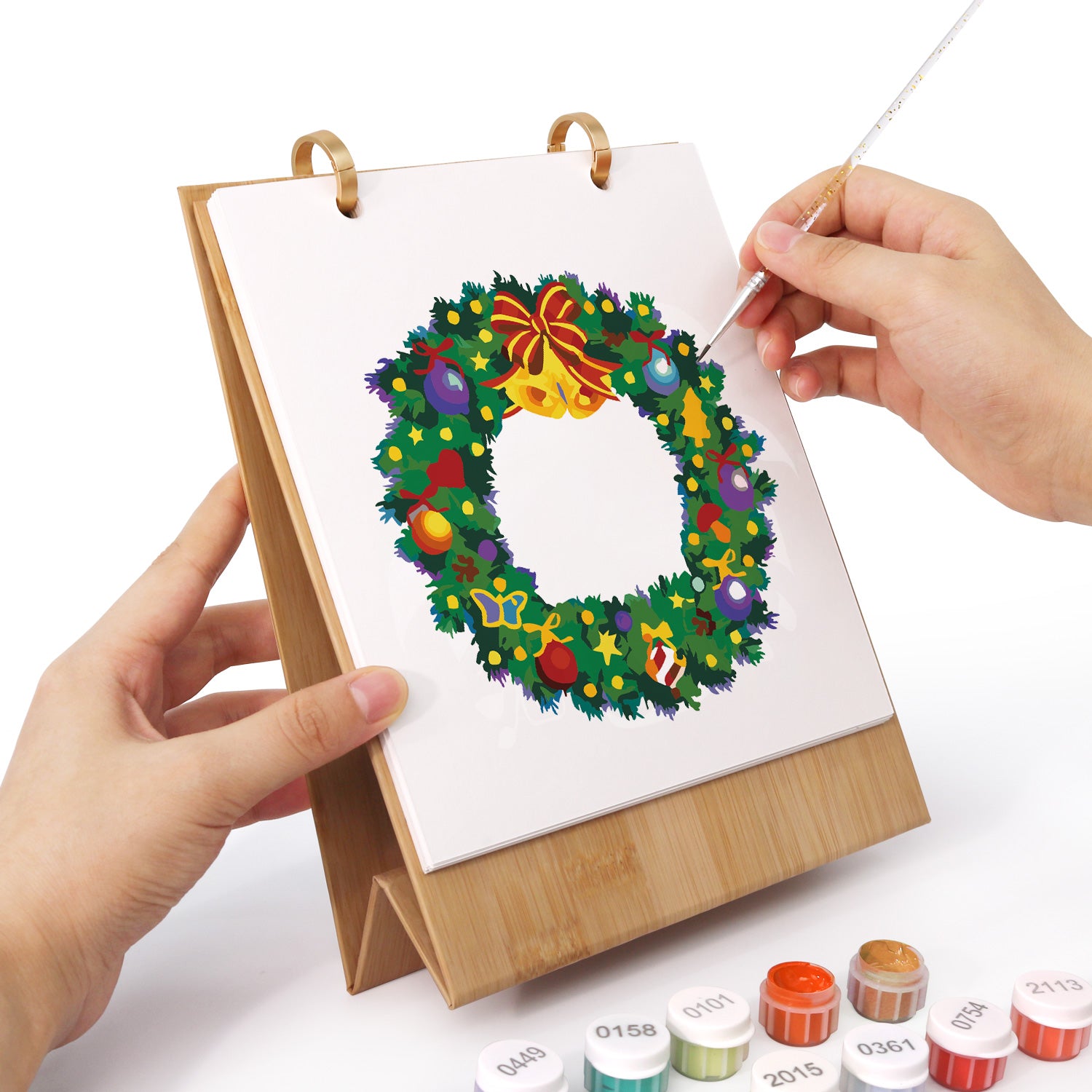 Person painting a Christmas wreath on a small easel with paint supplies.