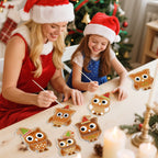 Woman and young girl wearing Santa hats painting owl ornaments on a table with a Christmas tree in the background.