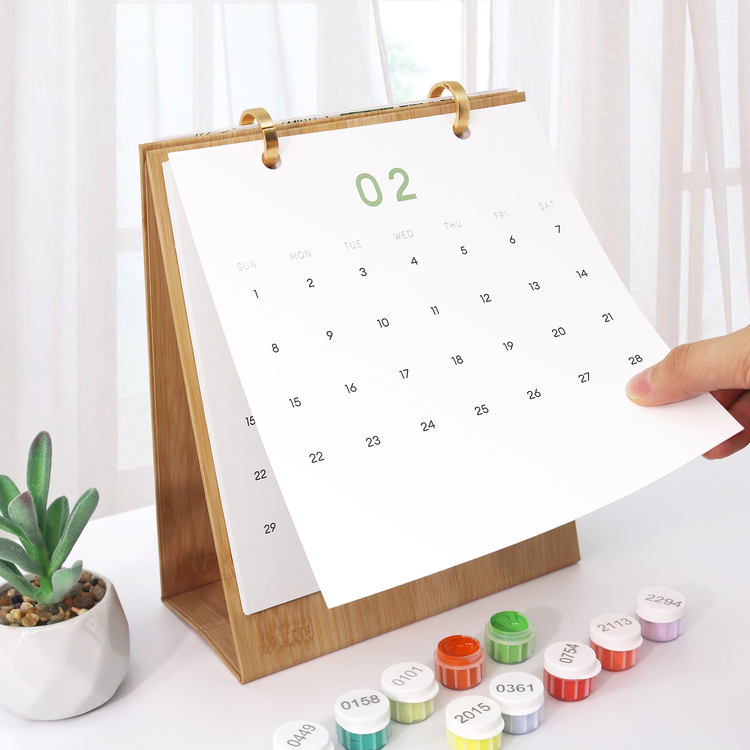 Calendario con soporte de madera sobre una superficie blanca con pequeños contenedores y una planta.