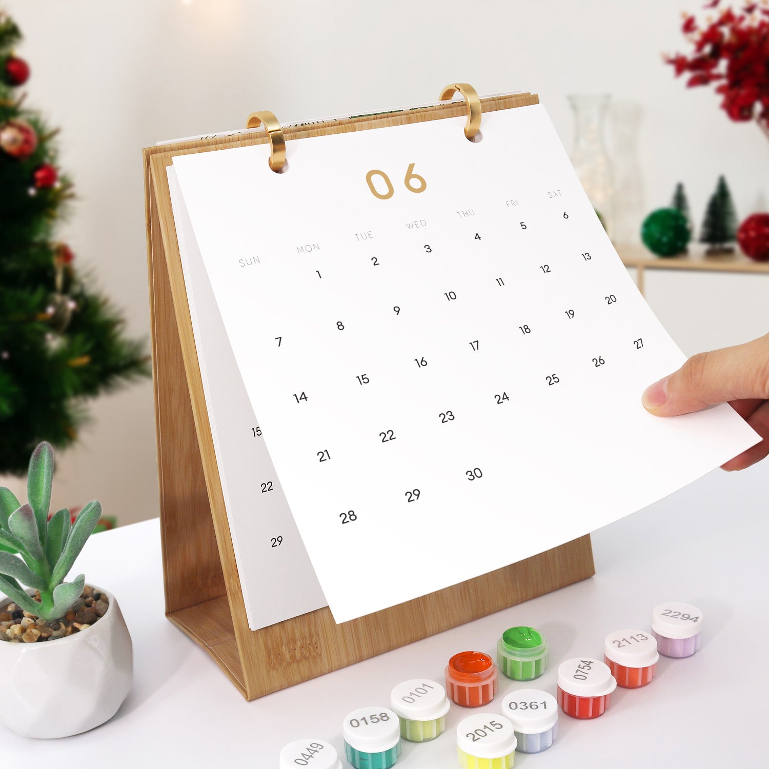 Calendario con soporte de madera sobre una mesa junto a pequeños contenedores, con un árbol de Navidad al fondo.