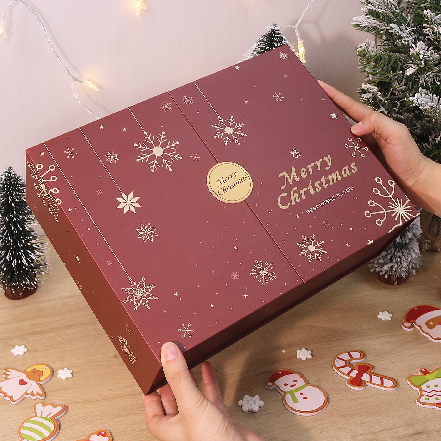 Red Christmas box with snowflakes and 'Merry Christmas' text, held by a hand on a wooden surface with festive decorations.