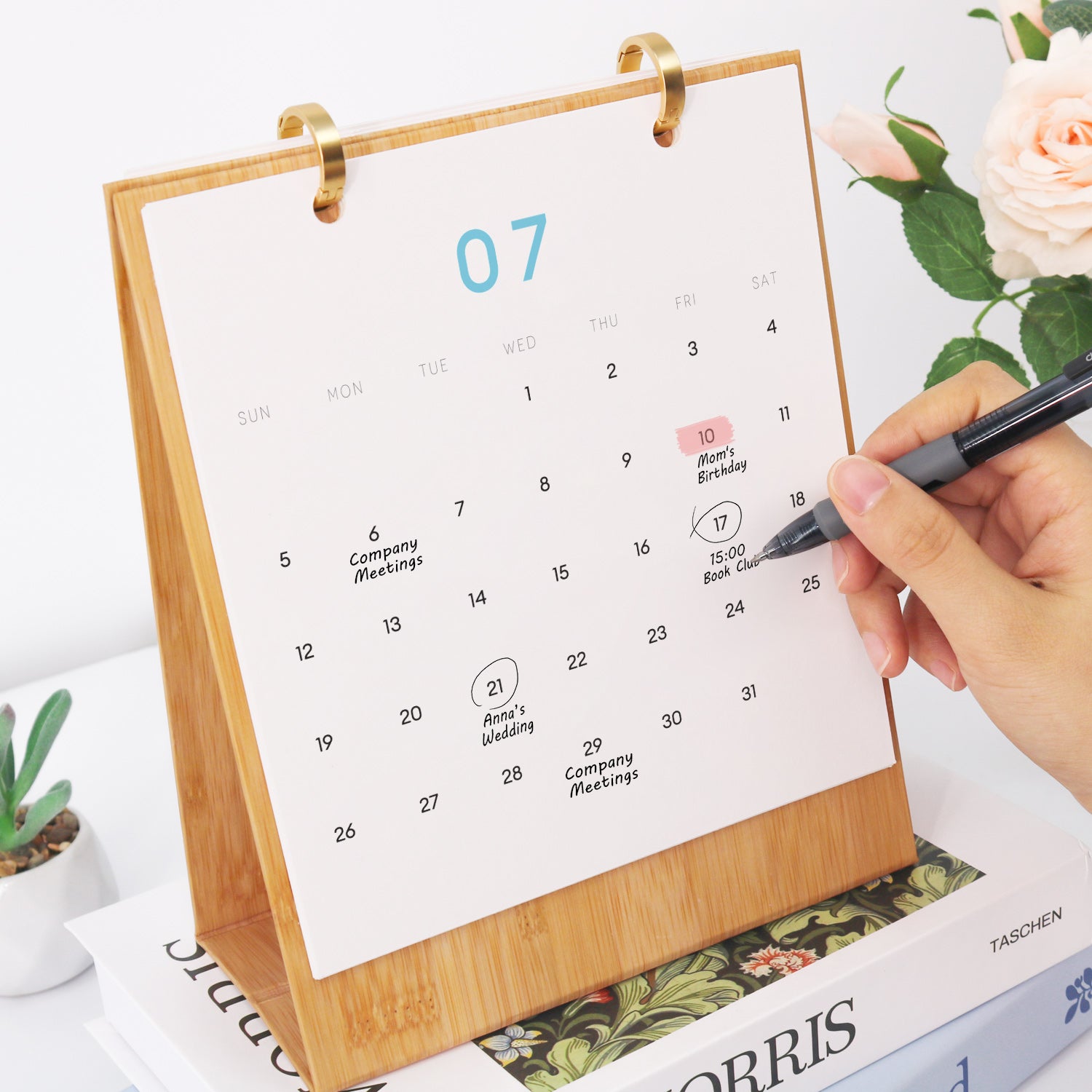 Kalender mit Holzständer auf einer Zeitschrift, Hand mit Stift und Blumen im Hintergrund