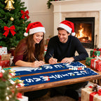 Two people wearing Santa hats sitting at a table with Christmas decorations and a fireplace in the background.