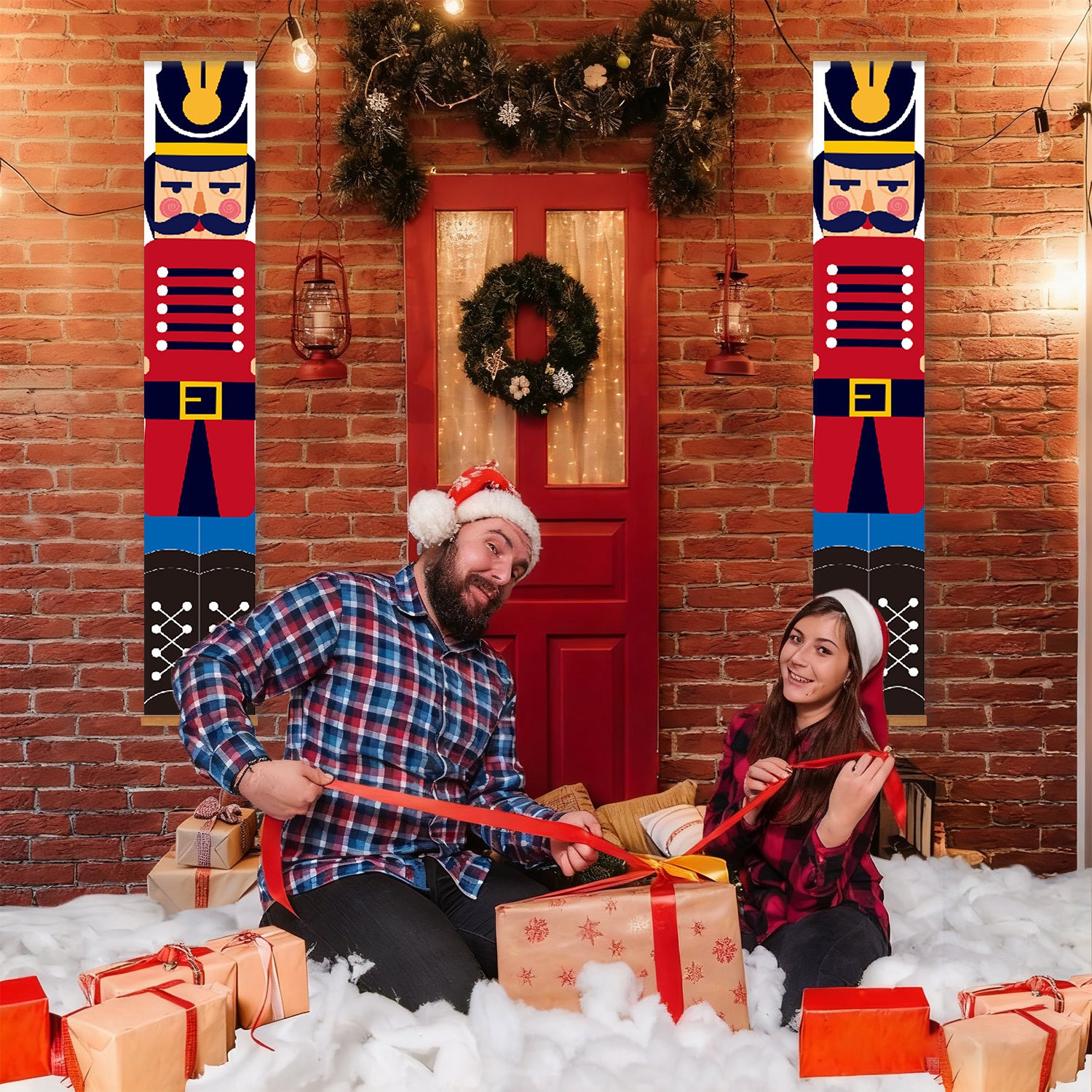 Two people sitting on a snowy floor with Christmas presents, wearing festive hats, in front of a red door with a wreath.