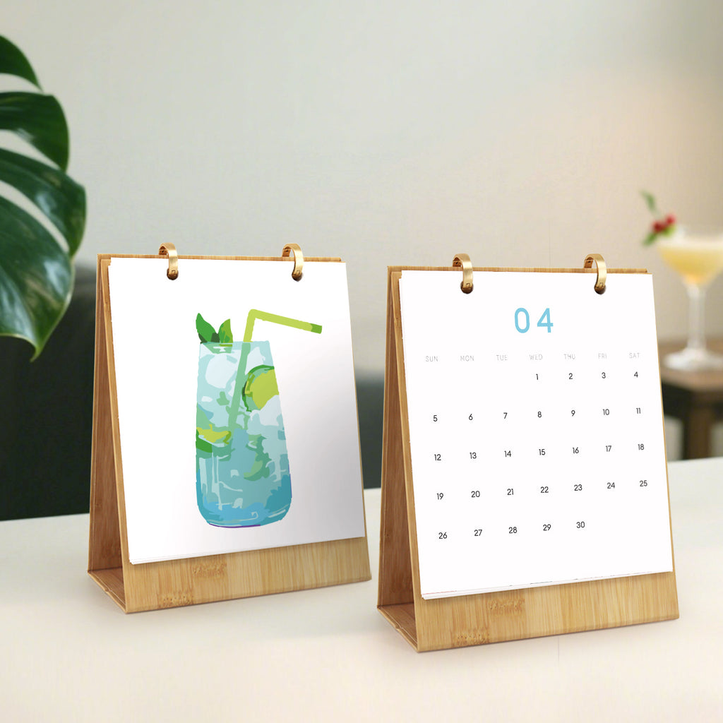 Dos pequeños calendarios de escritorio sobre una superficie con una planta y un cristal al fondo.