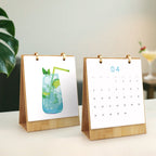 Two small desk calendars on a surface with a plant and glass in the background.