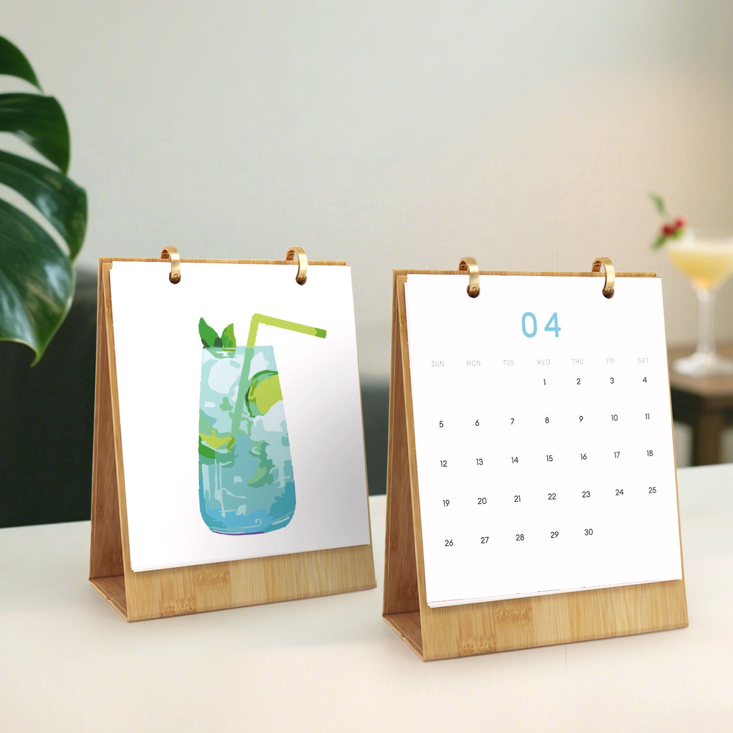 Two small desk calendars on a surface with a plant and glass in the background.