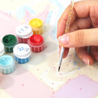 Hand holding a small brush over a piece of fabric with colorful paint containers in the background.