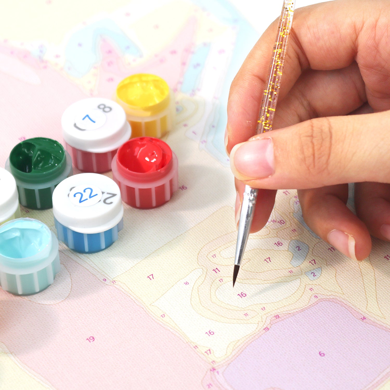 Hand holding a small brush over a piece of fabric with colorful paint containers in the background.