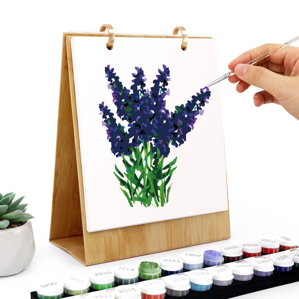 Hand painting a lavender flower on a white canvas with a wooden easel, surrounded by watercolor paints.