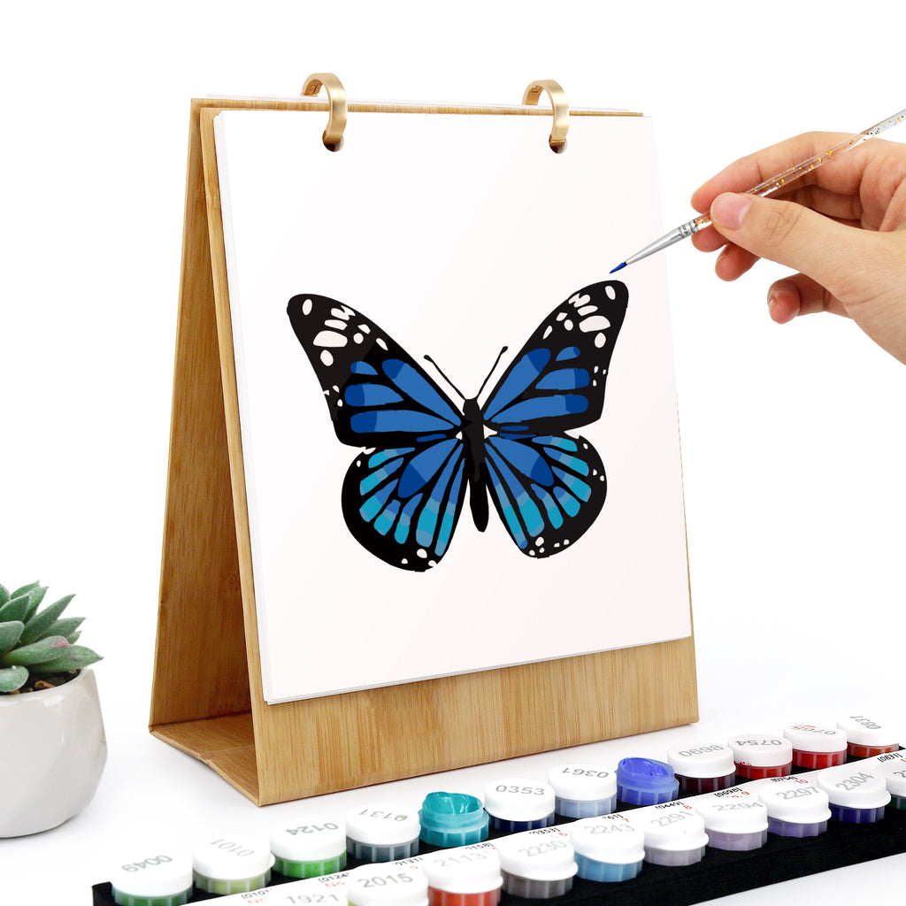 Person malt mit Wasserfarben einen blauen Schmetterling auf eine weiße Leinwand.