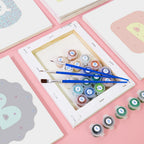 Set of paint tubes and brushes on a pink surface with a framed picture in the background