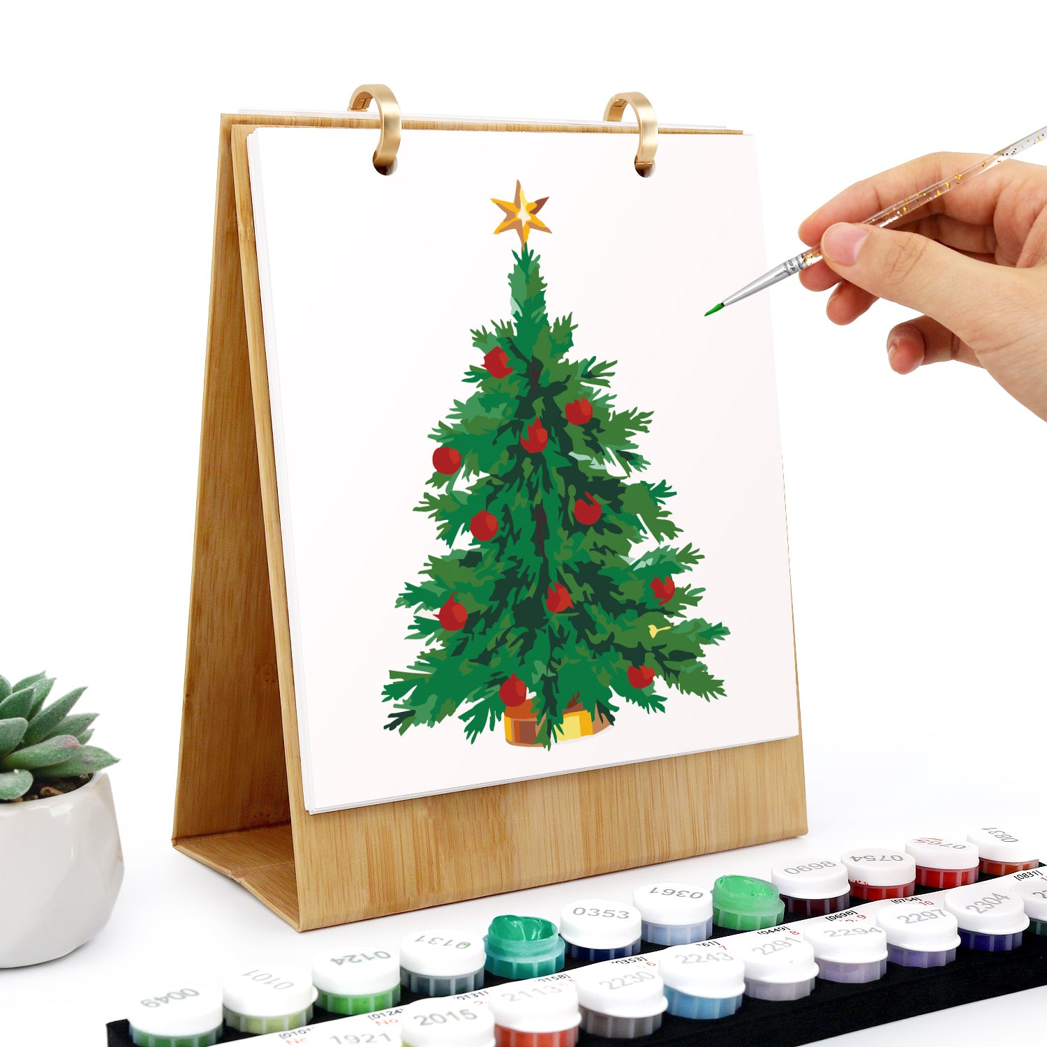 Hand painting a Christmas tree on a small canvas with watercolors and a paintbrush.