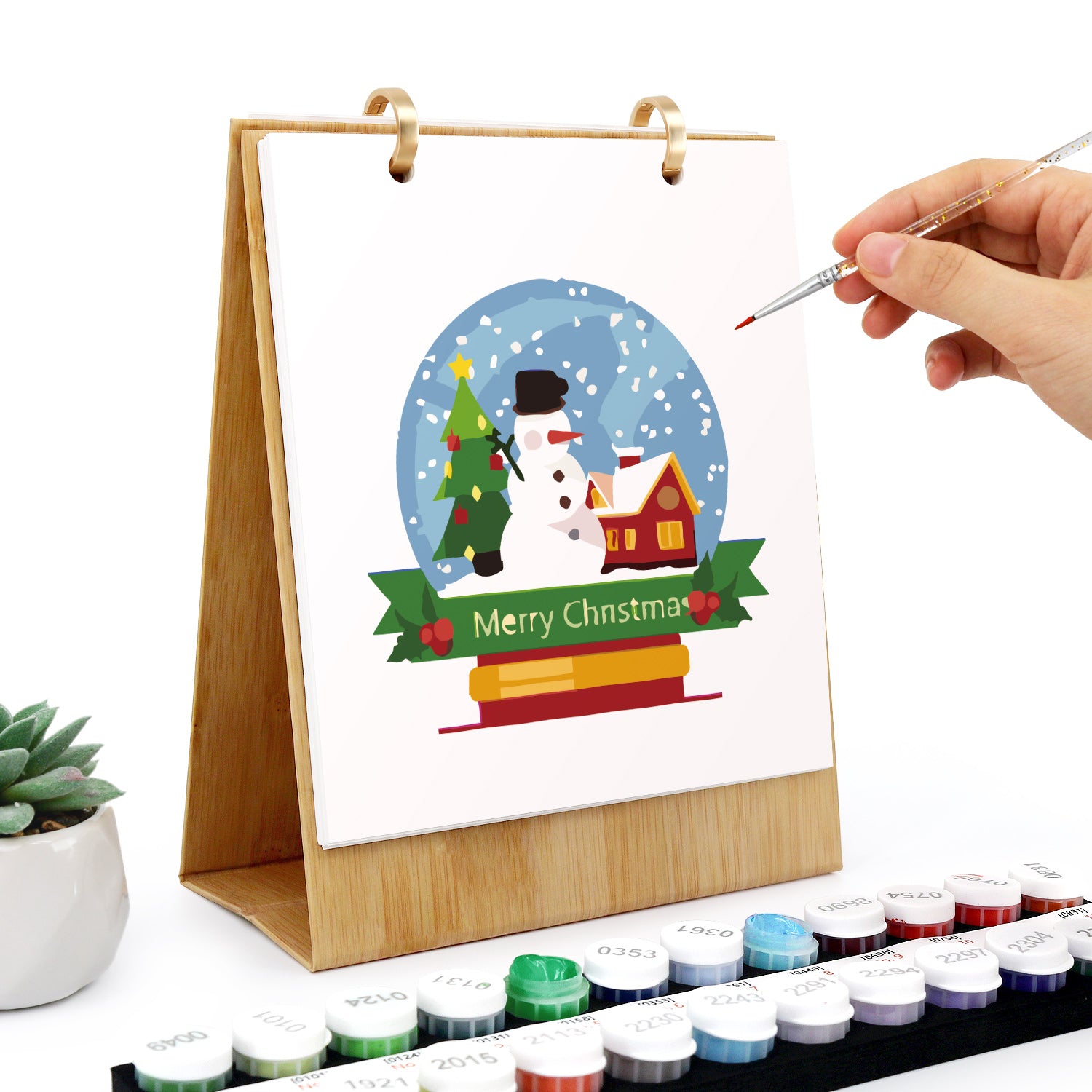 Hand painting a Christmas-themed scene on a white paper with a wooden easel and watercolor paints.