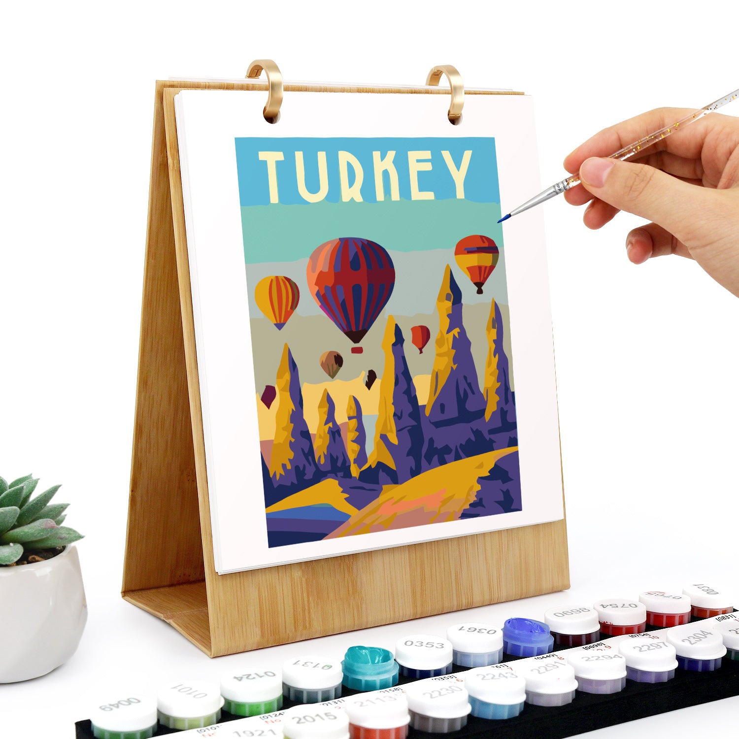 Colorful painting of Turkey with hot air balloons on a wooden easel, surrounded by watercolor paints.