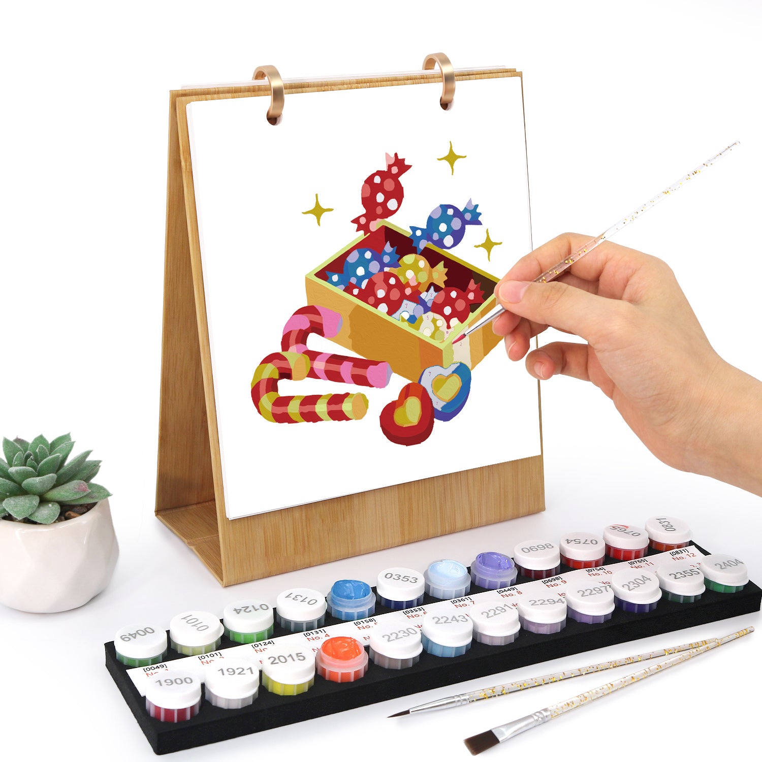 Configuración artística con pinturas de acuarela, pinceles y una mano pintando un diseño colorido sobre una superficie clara.