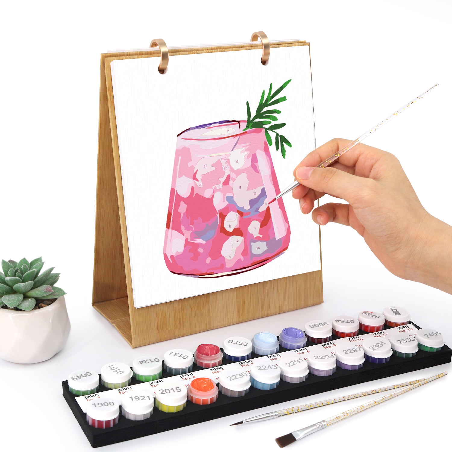 Handbemalung eines rosafarbenen Glases mit Blumenmuster auf einem Ständer, mit Aquarellfarben und Pinseln auf einer weißen Oberfläche.