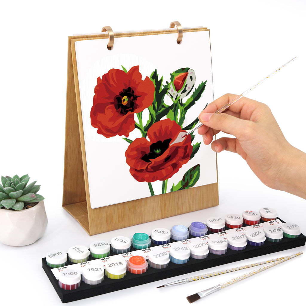 Person painting red flowers on a small easel with watercolor paints and brushes.