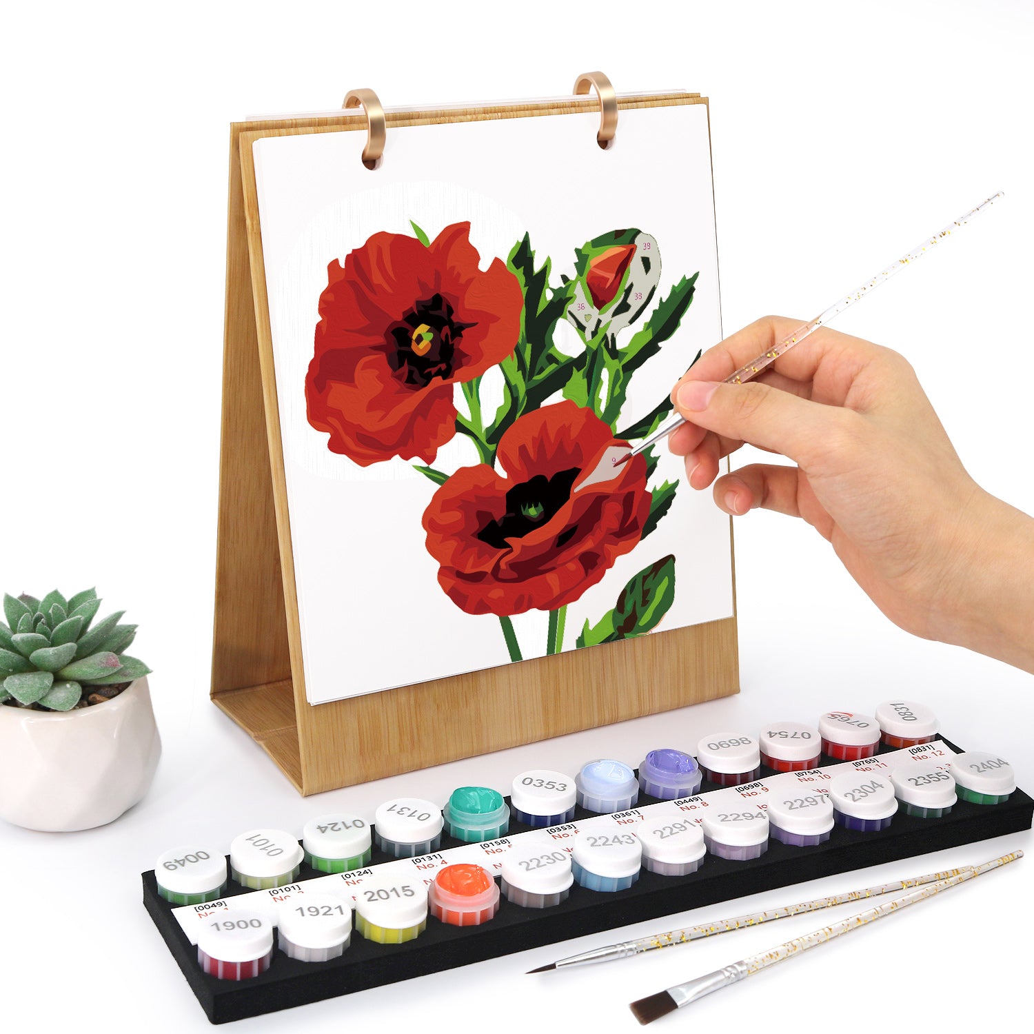 Person painting red flowers on a small easel with watercolor paints and brushes.