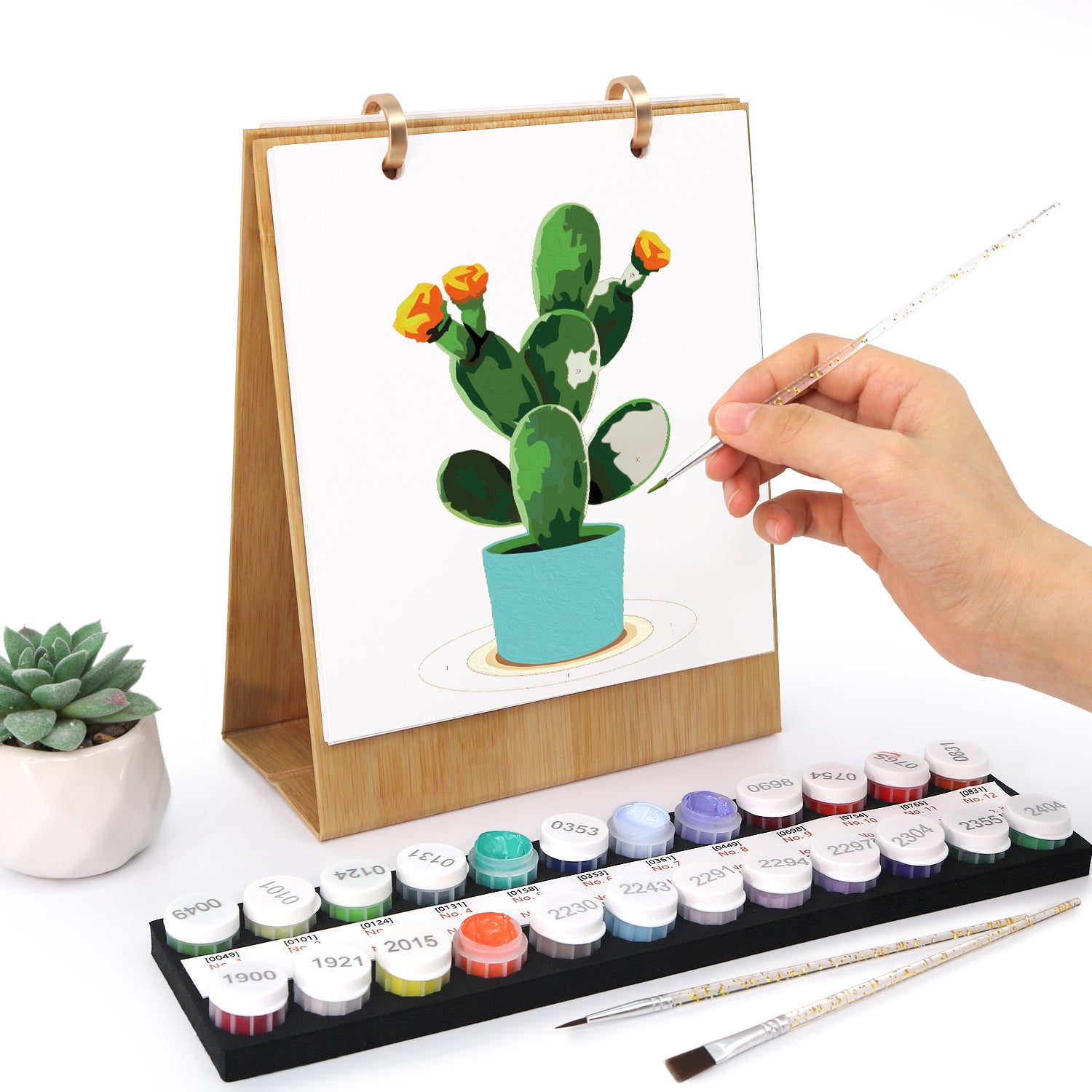 Person painting a cactus with watercolors on a small easel, surrounded by paint bottles and brushes.