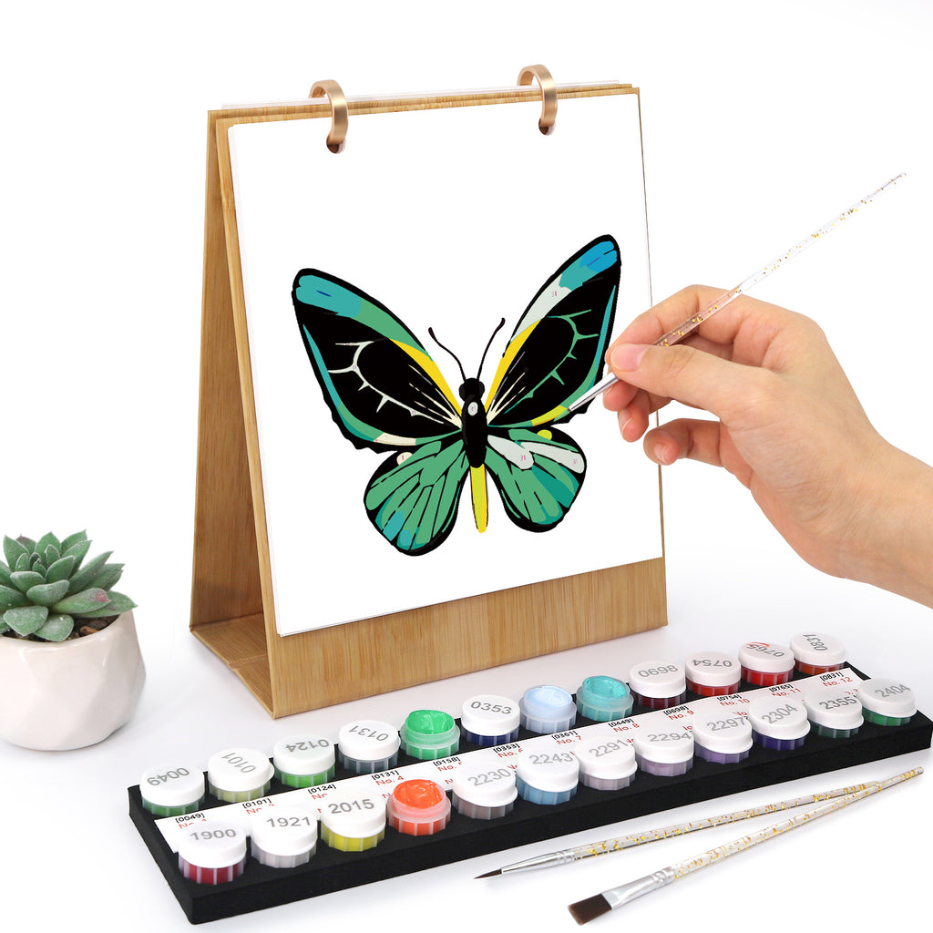 Person malt einen Schmetterling mit Wasserfarben auf einer weißen Oberfläche, umgeben von Farbflaschen und Pinseln.
