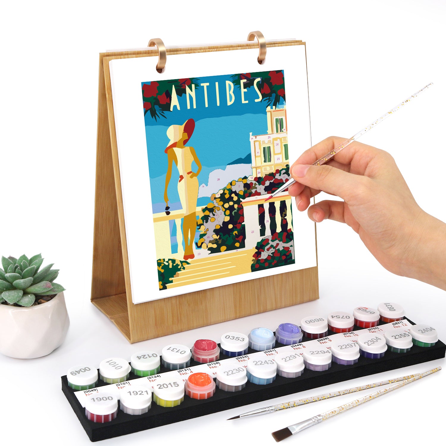 Artistic depiction of a scenic view with 'Antibes' text, held by a hand with a paintbrush, on a stand with watercolor paints and brushes.