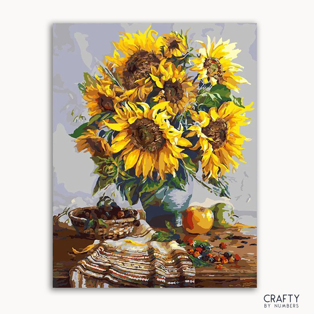 A paint-by-numbers kit image featuring a vase with vibrant sunflowers, alongside fruits and a cloth, set against a kitchen backdrop.