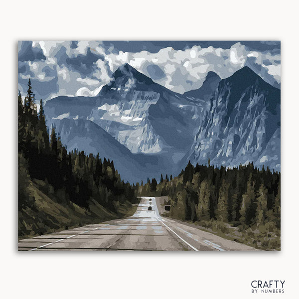 A framed canvas painting depicting a road leading towards a mountain range with a blue sky and clouds above.