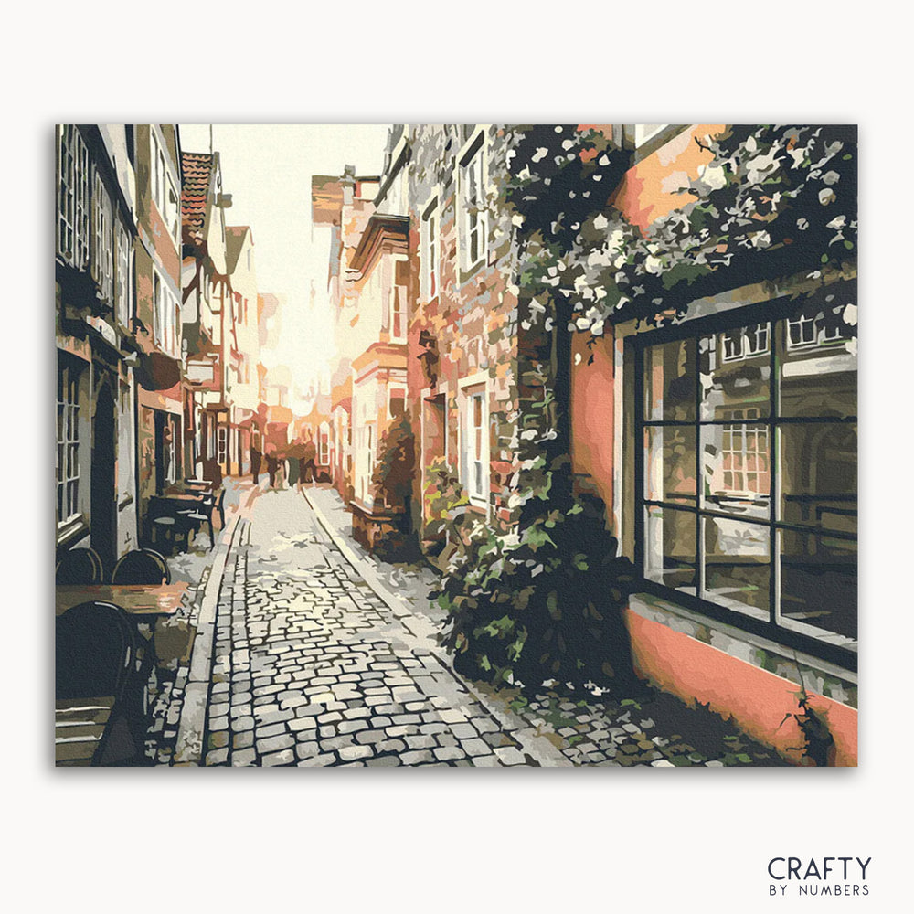 A canvas painting depicting a quiet street in an old town at sunset, with cobblestone pavement, buildings on either side, and hanging flowers.