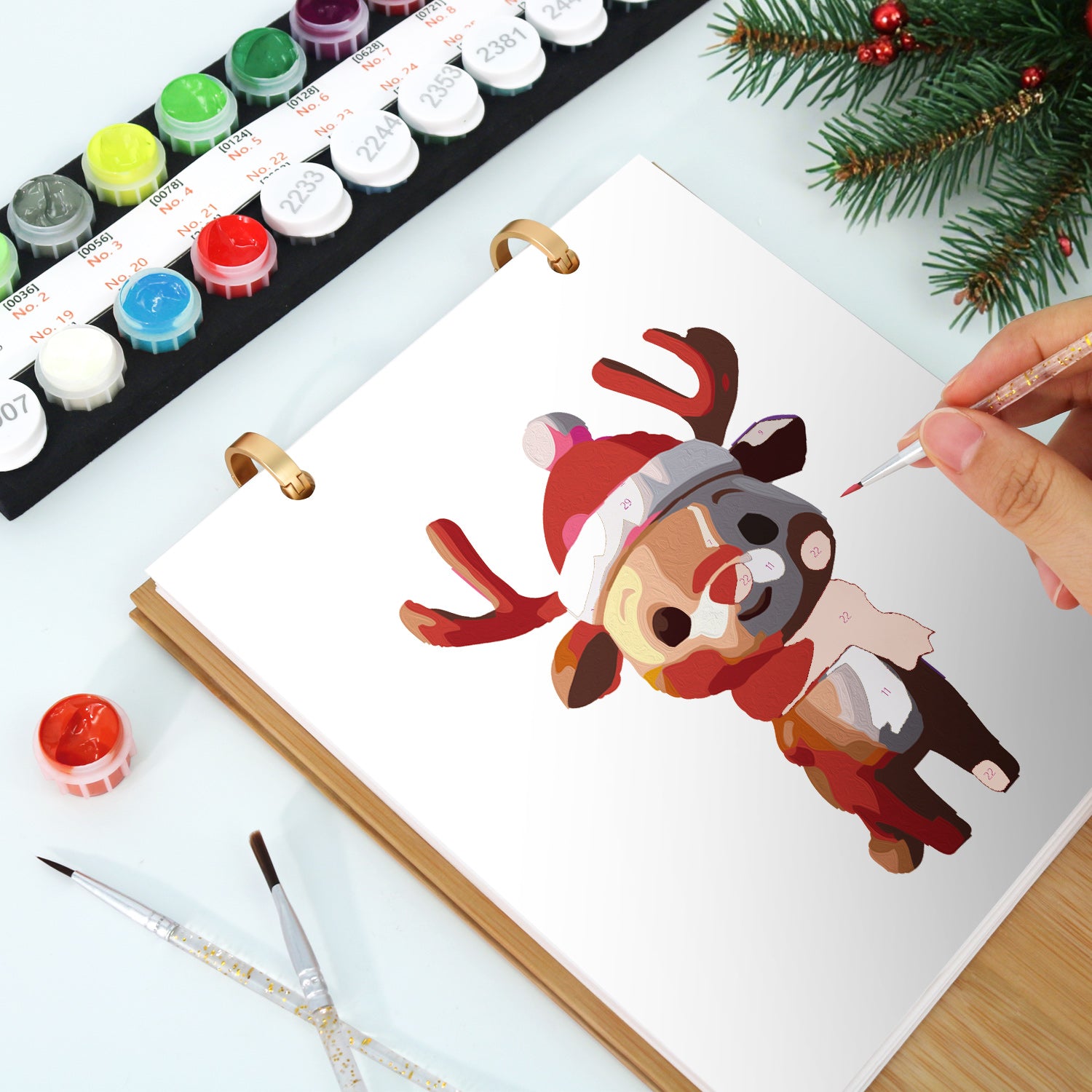 Person painting a cartoon reindeer with watercolors on a white paper, surrounded by art supplies and Christmas decorations.