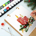 Person painting a Christmas-themed illustration of candles and holly leaves on a white sheet with watercolor paints and brushes.