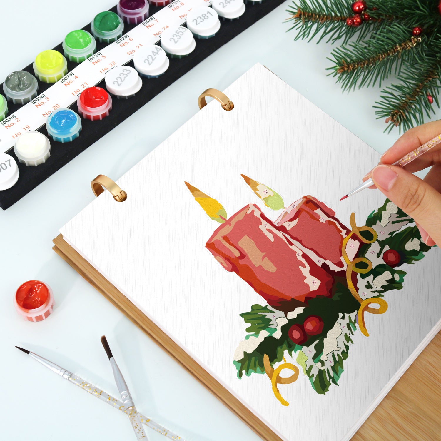 Person painting a Christmas-themed illustration of candles and holly leaves on a white sheet with watercolor paints and brushes.