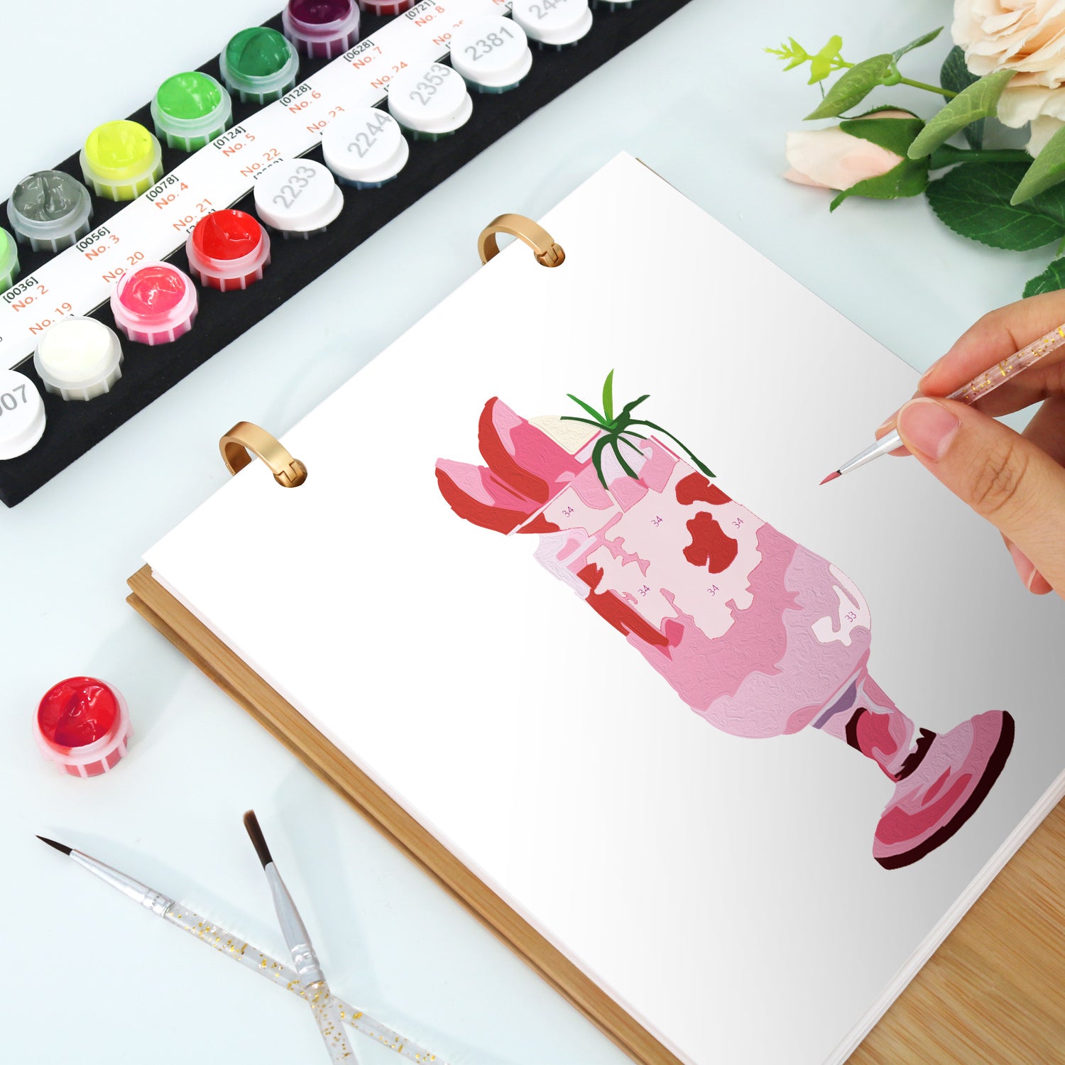 Person painting a pink glass with a straw on a white sheet of paper, surrounded by watercolor paints and brushes.