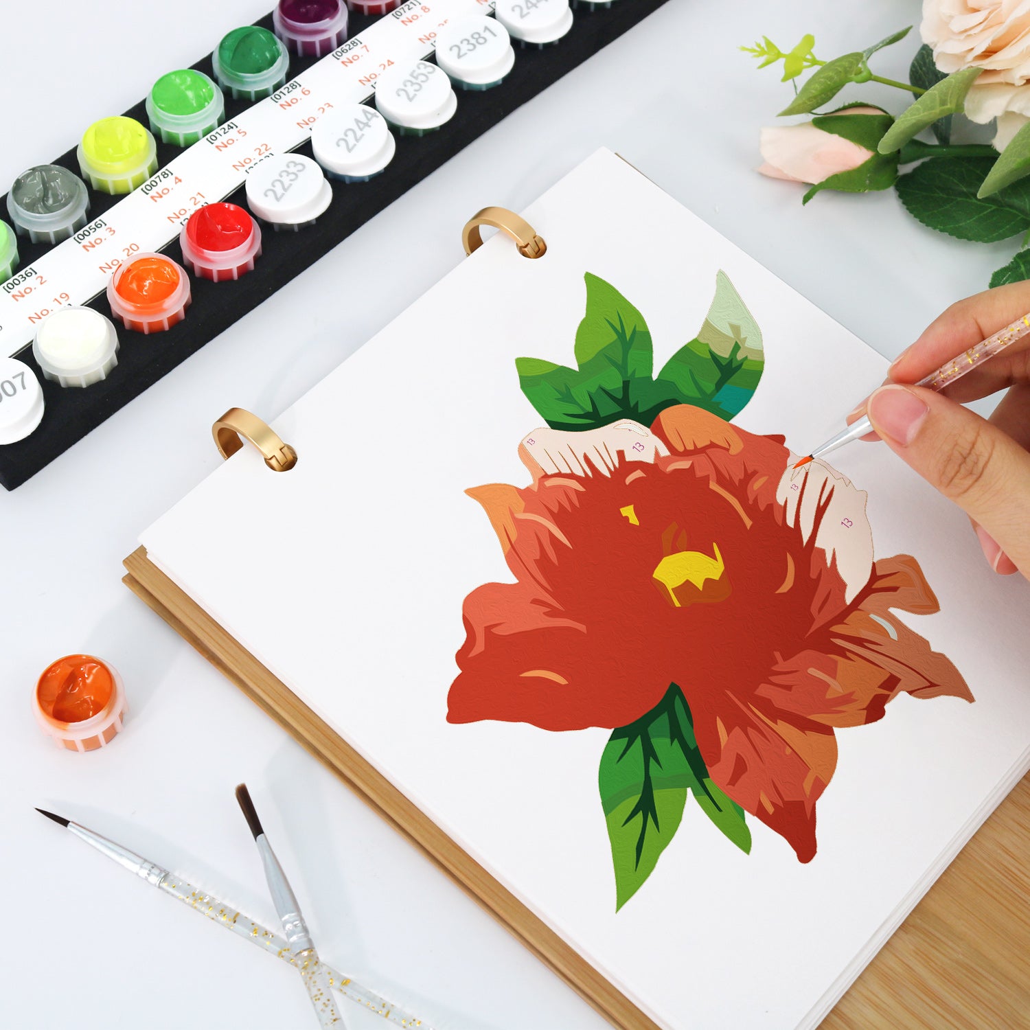 Person painting a red flower with green leaves on a white canvas, surrounded by art supplies.