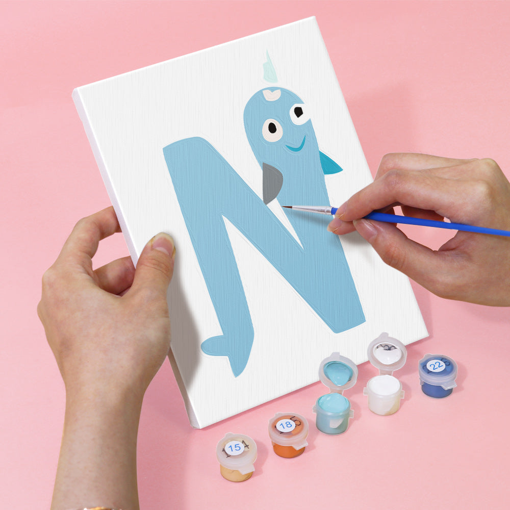 Person painting a blue letter 'N' with a creature on a white card against a pink background