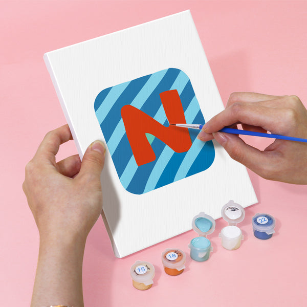 Person painting a red letter 'N' on a blue striped card with paint supplies on a pink background