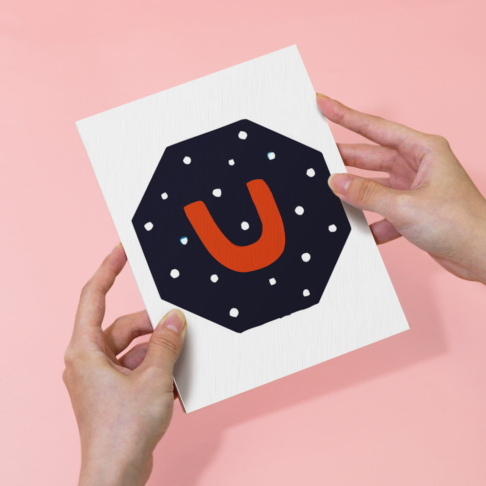 Card with letter 'U' design held by hands against a pink background