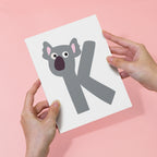 Card with a koala illustration and letter 'K' held by hands on a pink background