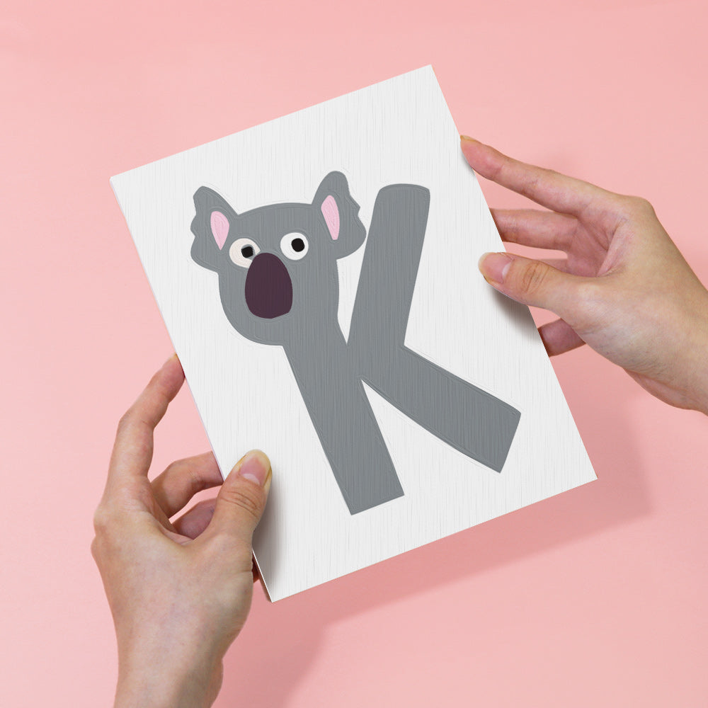 Card with a koala illustration and letter 'K' held by hands on a pink background