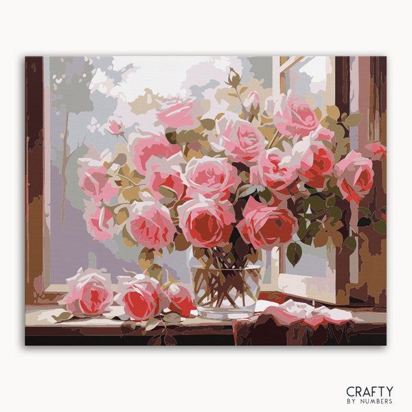A paint by numbers kit depicting a vase of pink roses placed on a windowsill, with the brand 'Crafty By Numbers' visible on the bottom right.