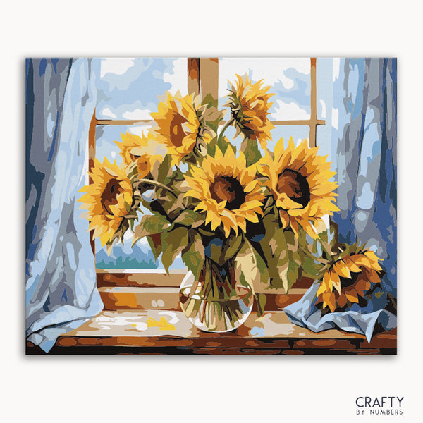 A paint by numbers kit depicting a vase of sunflowers placed on a windowsill, with a blue curtain and a bright window in the background.
