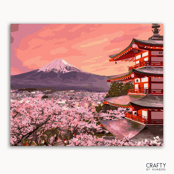 A framed canvas paint-by-numbers kit depicting a vibrant scene with Mount Fuji and a traditional Japanese structure surrounded by pink flowers at sunset.