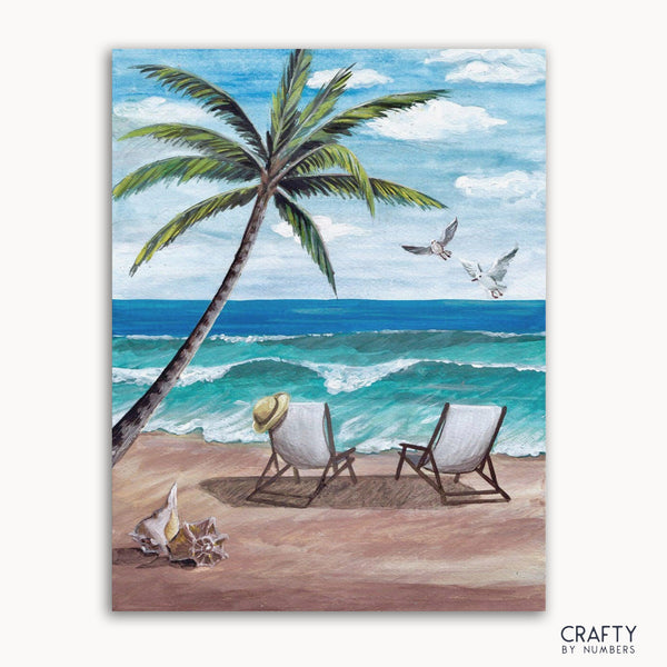 A paint by numbers kit depicting a beach scene with white sand, sunloungers, palm trees, and ocean waves.